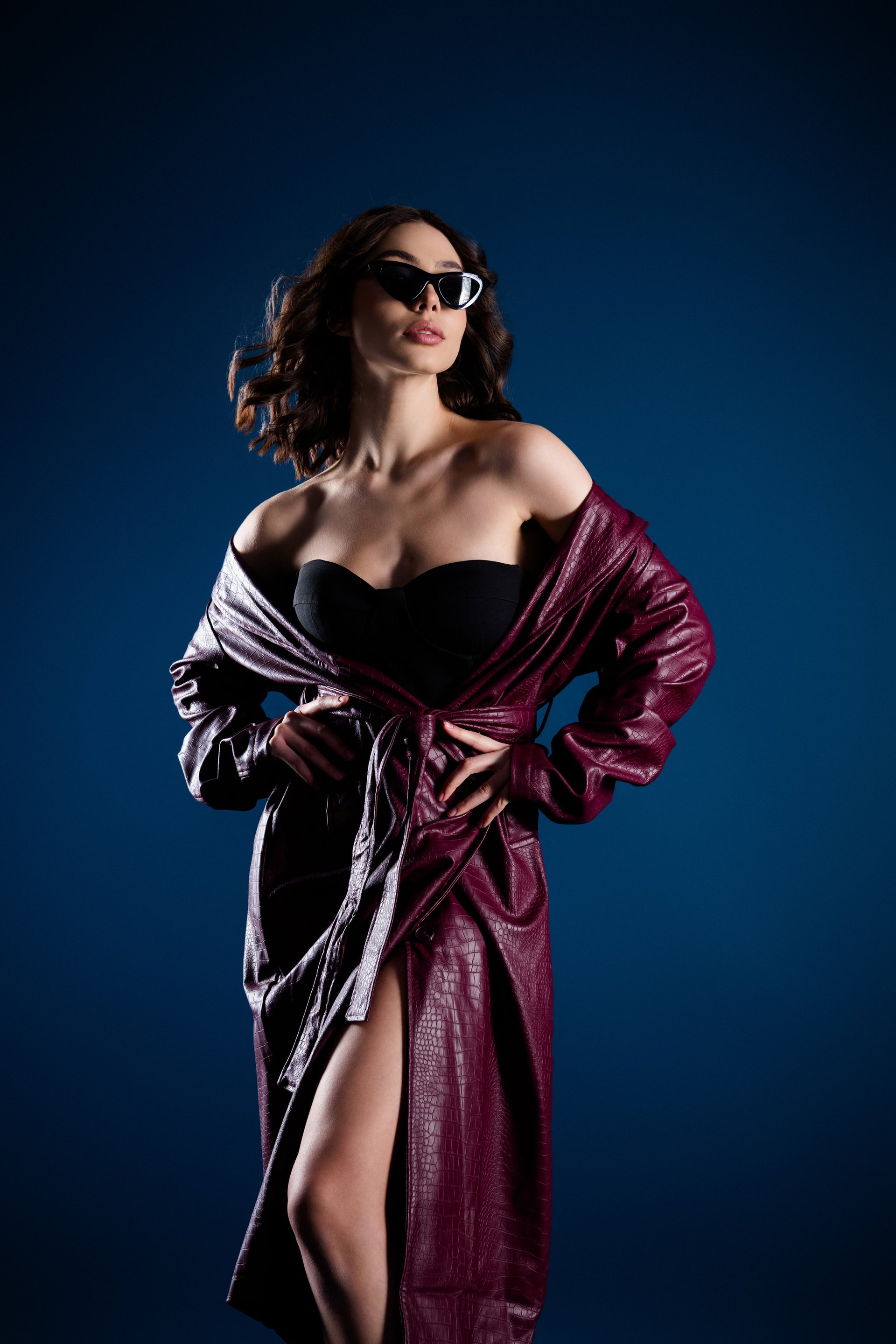 Fashion model poses confidently in a maroon leather coat and black strapless top, wearing sunglasses for a glamorous, stylish studio portrait that evokes luxury, attitude and evening chic.